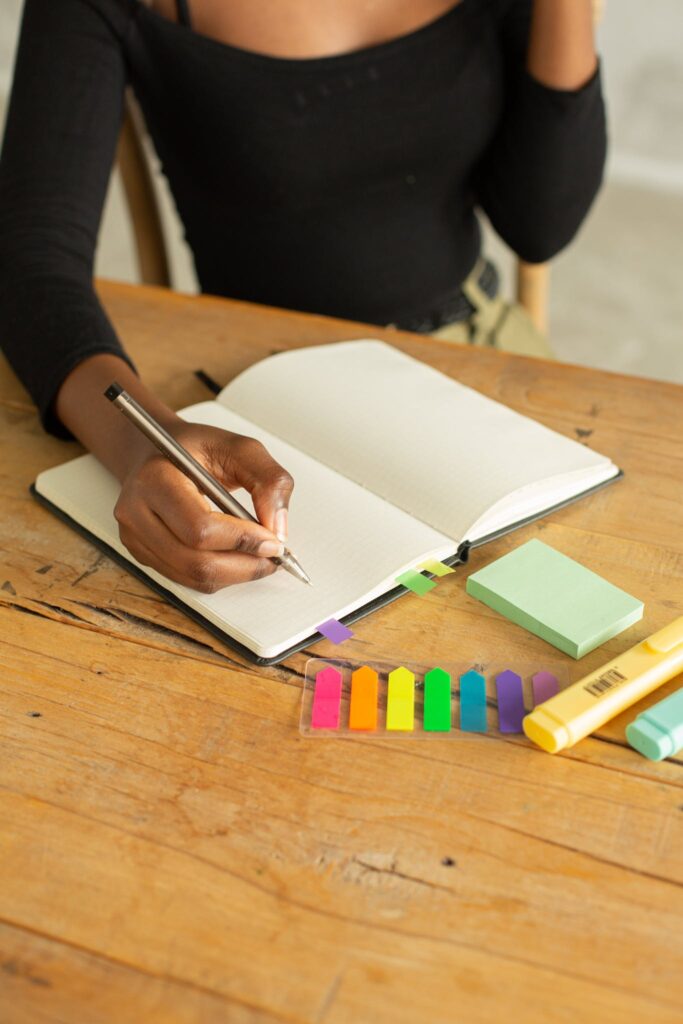 Crop unrecognizable ethnic female sitting at wooden table and taking notes on white blank page of notepad with pencil and markers